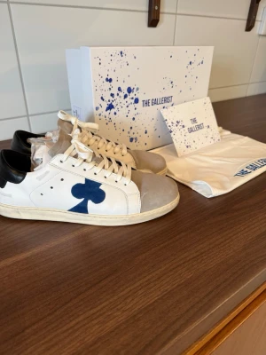 Vita sneakers från The Gallerist - Snygga vita sneakers från The Gallerist med blå klubb-symbol på sidan, svart hälparti och grå mockadetaljer vid tån. Skorna har vita snören och en klassisk låg siluett. Perfekta för dig som vill sticka ut med en unik design och coola detaljer. Skorna är använda 1 gång så inprinsip helt nya.