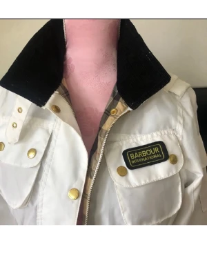 Vit Barbour jacka med svarta detaljer - Snygg vit jacka från Barbour International med svart krage, guldiga knappar och flera fickor framtill. Klassisk patch på bröstet och rutigt innerfoder. Jackan har dragkedja och tryckknappar för extra stilpoäng.