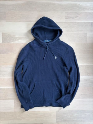 Mörkblå hoodie från Polo Ralph Lauren - Säljer en mörkblå hoodie från Polo Ralph Lauren med klassisk huva och dragsko. Tröjan är ribbstickad och har en broderad vit logga på bröstet. Perfekt för en avslappnad och stilren look. Känguruficka framtill och långa ärmar.