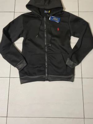 Svart hoodie från Polo Ralph Lauren - Svart hoodie med dragkedja från Polo Ralph Lauren. Klassisk design med huva och dragsko, samt röd broderad logga på bröstet. Tillverkad i mjukt material och har fickor framtill. Perfekt för en avslappnad och stilren look.