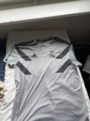 Grå Real Madrid träningströja Adidas - Snygg grå träningströja från Adidas med Real Madrid-logga på bröstet. Tröjan har svarta detaljer och ränder på axlarna samt grafiskt mönster framtill. Tillverkad i ett lätt och funktionellt material som passar perfekt till träning.