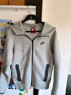Nike tech fleece  - Snygg grå hoodie från Nike med dragkedja framtill och två svarta dragkedjeförsedda fickor. Klassisk Nike-logga på bröstet och huva baktill. Tillverkad i mjukt material som känns skönt mot huden. Perfekt för en avslappnad och sportig stil. Tyvärr lite deffekt dragkedja nertill men går att stänga o öppna 