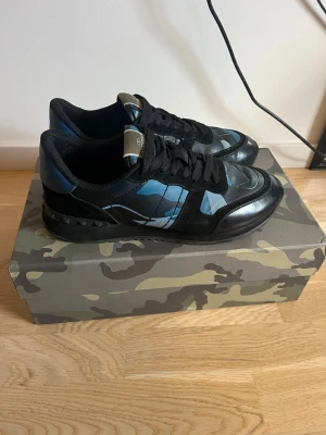 Valentino blå camo sneakers - Snygga sneakers från Valentino med blå och svart camo-mönster. Skorna har svarta snören, svarta detaljer i läder och textil samt en unik sula med nitar baktill. Perfekta för dig som vill sticka ut med en cool och modern stil. St 42 men passar även 43 pris kan diskuteras