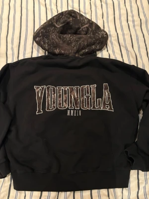 Svart hoodie från YoungLA med tryck - Svart hoodie från YoungLA med stort tryck 'YOUNGLA MMXIV' på ryggen och mindre logga på fickan framtill. Huvan har ett unikt mönster i mörka toner och tröjan har en klassisk magficka. Perfekt för en avslappnad streetwear-look.