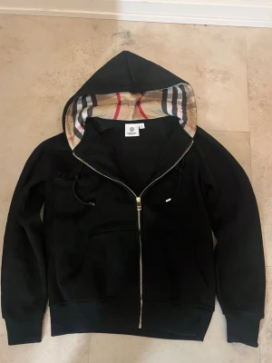Svart hoodie med dragkedja och mönstrad huva - Svart hoodie med hel dragkedja och klassisk mönstrad insida på huvan i beige, rött, blått och vitt. Hoodien har långa ärmar, snörning i huvan och fickor framtill. Perfekt för en avslappnad och stilren look.