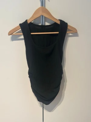 Svart ribbad linnetopp - Snygg svart ribbad linnetopp med rund halsringning och åtsittande passform. Toppen är ärmlös och har en lätt rynkad detalj i nederkanten som ger en cool twist. Perfekt att styla med jeans eller kjol för en trendig look.