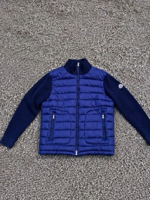 Moncler Cardigan  - Snygg mörkblå dunväst med quiltad framsida och stickade ärmar. Två dragkedjeförsedda fickor framtill och hög krage. Materialmix av dun och stickat tyg ger en cool och modern look. Perfekt för lager-på-lager och streetstyle.