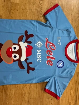 Napoli jultröja med renmotiv - Ljusblå Napoli jultröja från EA7 med stort renmotiv på framsidan, röda detaljer vid ärmslut och krage samt tryckta loggor som 'Lete', 'MSC' och Napoli-emblem. Tillverkad i ett lätt och sportigt material, perfekt för dig som vill sticka ut på match eller julfest.     Fraktar samma eler dagen efter du köpt!!! Endast en kvar i lager!!!