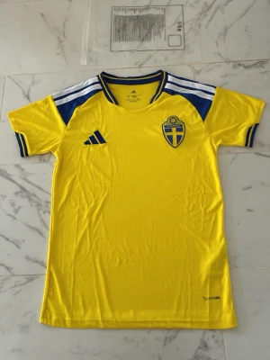 Svensk gul fotbollströja Adidas - Gul fotbollströja från Adidas med blå detaljer och Sveriges landslagsemblem på bröstet. Tröjan har korta ärmar, rund hals och klassiska Adidas-ränder på axlarna. Tillverkad i lätt och ventilerande material, perfekt för träning eller match. Pris kan diskuteras vid snabb affär!