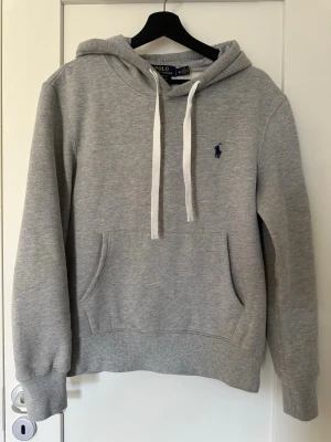 Grå hoodie från Polo Ralph Lauren -  Sitter bra om man vanligtvis har S i kläder.Snygg grå hoodie från Polo Ralph Lauren med vit dragsko, klassisk känguruficka och broderad marinblå logga på.