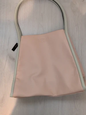 Ljusrosa shoppingväska med vita detaljer - Snygg ljusrosa shoppingväska från Victoria's Secret med vita kanter och långa handtag. Väskan har en enkel och stilren design i ett mjukt material, perfekt för att bära med sig allt man behöver. Märkesloggan är diskret präglad på framsidan.