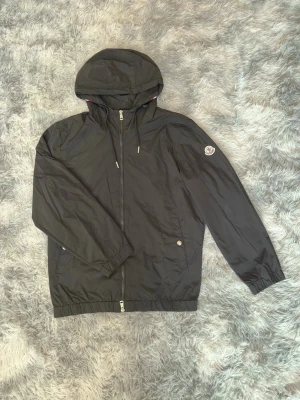 Moncler Windbreaker  - Snygg svart vindjacka med huva och dragkedja framtill. Jackan har två fickor med knapp, elastiska muddar och en patch på ärmen. Huvan har dragsko och en rödvit rand baktill. Perfekt för blåsiga dagar och enkel att matcha.
