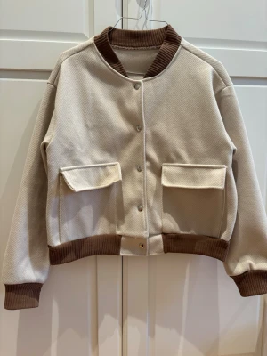 Beige bomberjacka med bruna detaljer - Snygg beige bomberjacka med bruna ribbade muddar och krage. Köpt second hand men är i ett mycket bra skick!