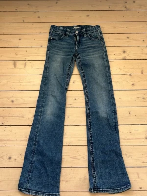 Blå bootcut jeans med kontrastsömmar - Snygga blå bootcut jeans med slitningar och markerade kontrastsömmar. Jeansen har två bakfickor med lock och silverfärgade knappar. Klassisk femficksmodell i denim med lätt tvättad look och utsvängda ben.