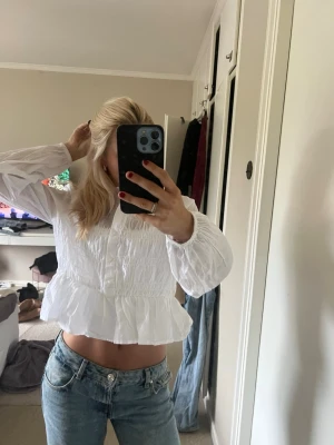 Vit volangblus med lång ärm - Säljer en söt vit blus med volang nertill och smockad midja. Blusen har lång ärm med resår vid ärmslut och en lätt krinklad struktur i tyget. Perfekt att styla med jeans för en trendig look.