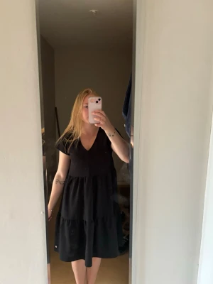 Svart v-ringad kortklänning - En svart kortklänning med v-ringning och volangdetaljer. Klänningen har korta ärmar och en lös passform som ger en avslappnad look. Perfekt för dig som gillar enkel men snygg stil.