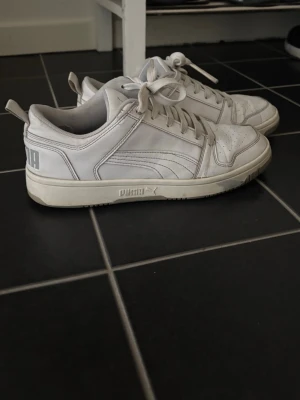 Vita låga sneakers från Puma - Säljer ett par vita sneakers från Puma med klassisk låg siluett. Skorna har perforerad tå, vita snören och diskret Puma-logga på sidan och hälen. Tillverkade i slätt lädermaterial för en clean och stilren look.