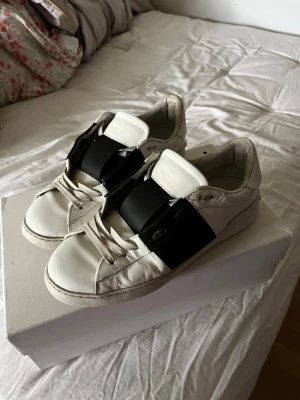 Valentino vita sneakers med svart detalj - Säljer ett par vita sneakers från Valentino med bred svart läderdetalj över snörningen. Skorna har klassisk låg profil, rund tå och platt sula. Tillverkade i läder med diskret logga på plösen. Perfekta för dig som gillar stilrena men ändå unika sneakers.