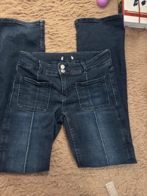 Mörkblå bootcut jeans med fickor - Säljer ett par mörkblå bootcut jeans med dubbla knappar i midjan och stora framfickor. Jeansen har markerade sömmar och klassiska bakfickor. Materialet är denim med lätt tvättad look och raka ben som går ut något nedtill. materialet är väldigt stretchig. Strl 164 fast sitter som xs-s.  Midjemått:35cm Innerbenslängd:76
