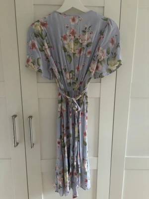 Ljusblå blommig långklänning med knyt - Söt ljuslila långklänning med blommigt mönster. Klänningen har v-ringning, korta ärmar och ett knytband i midjan för en smickrande passform. Perfekt för sommarens alla tillfällen!