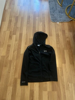 Svart träningsjacka från Under Armour - Svart träningsjacka med huva från Under Armour. Jackan har dragkedja framtill, två fickor och reflekterande detaljer vid axlarna. Tillverkad i lätt syntetmaterial, perfekt för utomhusaktiviteter.