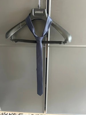 Mörkblå smal slips - En stilren, smal slips från J.Lindbergh i mörkblått tyg. Perfekt för dig som vill ha en clean och klassisk look till skjortan. Slipsen har en matt finish och är enkel att matcha med olika outfits.