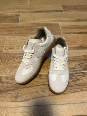 Beige sneakers med gummisula - Snygga beige sneakers med vit panel och detaljer i mocka och textil. Skorna har klassisk låg profil, vita skosnören och en brun gummisula som ger en retrovibe. Perfekta för en clean och stilren look.De är i otroligt bra skick. De har aldrig använts på grund av storleksproblem. Och om de var i min storlek skulle jag aldrig sälja dem på grund av hur rena och minimalistiska de ser ut och hur de skulle passa ihop med allt, jeans, mjukisbyxor och shorts. Tyvärr har jag inte märka eller originala låda
