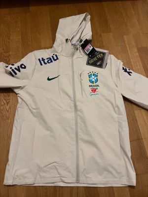 Brasilien Beige Windbreaker | Nike | Storlek L - Snygg beige vindjacka från Nike med Brasiliens landslagslogga på bröstet och blåa tryck på ärmarna. Jackan har huva, hel dragkedja och är tillverkad i GORE-TEX-material som står emot regn och vind. Perfekt för dig som gillar sportig stil och vill sticka ut. Perfekt för dina sommar outfits och matchar i princip allt. Storlek L