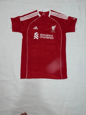 Liverpool röd matchtröja Adidas storlek M - Röd Liverpool FC matchtröja från Adidas med vita detaljer och klubbmärke på bröstet. Tröjan har korta ärmar, rund hals och vita ränder på axlarna. Tillverkad i lätt och ventilerande material, perfekt för fotboll eller träning.