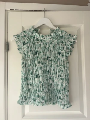 Grönblommig blus - Supersöt blus i vitt med grönt blommönster och plisserad struktur. Blusen har volangdetaljer vid hals och ärmslut samt korta ärmar. Säljer då den inte passar min stil längre💗Jätteliten gul fläck på ryggen som syns på bild 3