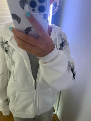 Vit hoodie med svart mönster - Säljer en vit hoodie med dragkedja och huva. Hoodien har svarta grafiska mönster på ärmar och axlar samt stora fickor framtill. Tillverkad i mjukt material, perfekt för en chill och avslappnad stil.                                      Köpt i Tyskland💗 Storlek M, men passar även S och L Skriv för flera bilder