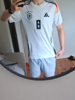 Tysklands fotbollströja Kroos #8 - Säljer en vit Tyskland fotbollströja med svarta detaljer och nummer 8 på bröstet och ryggen. Kroos står på ryggen. Tröjan har ränder i svart, rött och gult på axlarna och är tillverkad i ett lätt, andningsbart material. Perfekt för match eller träning.