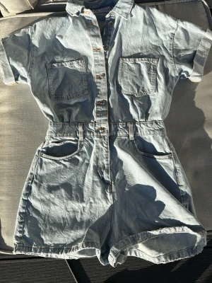 Ljusblå jeansjumpsuit med korta ärmar - Säljer en ljusblå jeansjumpsuit i denim med korta ärmar, krage och knappar framtill. Modellen har två bröstfickor, två bakfickor och två sidofickor samt resår i midjan för en snygg passform. Perfekt för en avslappnad och trendig look.