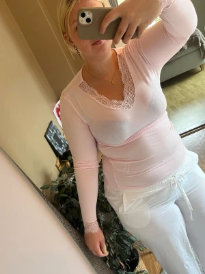 Ljusrosa loungewear-set med spets - Säljer ett ljusrosa loungewear-set med långärmad topp och vita byxor. Toppen har v-ringning med spetsdetaljer vid halsen och ärmsluten, samt en mjuk och stretchig passform. Byxorna är vita med dragsko i midjan för extra komfort. Perfekt för mysiga hemmakvällar.