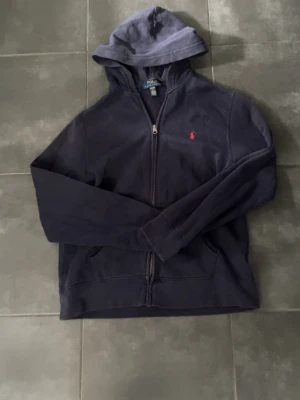 Mörkblå hoodie från Polo Ralph Lauren - Mörkblå hoodie med dragkedja från Polo Ralph Lauren. Klassisk design med huva, fickor framtill och den ikoniska röda loggan broderad på bröstet. Perfekt för en avslappnad och stilren look. Tillverkad i mjukt bomullsmaterial.