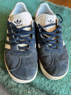 Adidas Gazelle mörkblå sneakers - Säljer ett par klassiska Adidas Gazelle sneakers i mörkblå mocka med vita detaljer och vita ränder på sidorna. Skorna har mörkblå snören och vit sula. Adidas-logga på plösen och sidan. Slitna bak på sulan men har mycket kvar att ge. Därav priset också. Skicka dm vid frågor. 