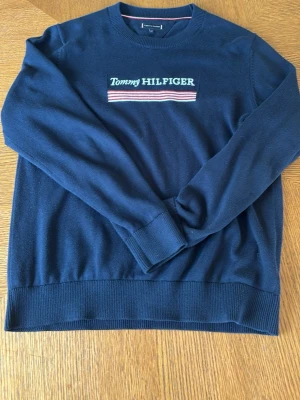 Marinblå stickad tröja Tommy Hilfiger - Snygg marinblå stickad tröja från Tommy Hilfiger med broderad logga och ränder i vitt, rött och blått på bröstet. Tröjan har rund halsringning, långa ärmar och ribbade muddar. Perfekt för dig som gillar klassisk och stilren design. 