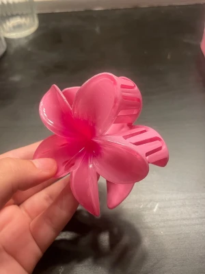 Rosa blomformad hårklämma - Säljer en söt hårklämma i plast formad som en blomma i en klar rosa färg. Perfekt för att piffa upp frisyren med en färgglad och lekfull detalj. Klämman har fem blomblad och är enkel att använda för att hålla håret på plats.
