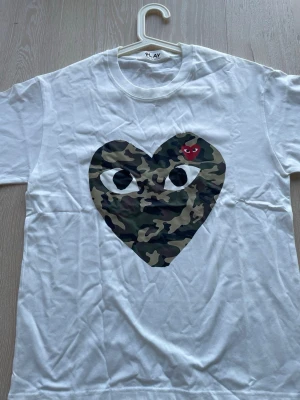 Vit t-shirt med camo hjärta - Säljer en vit t-shirt med ett stort hjärta i camouflage-mönster och ögon på bröstet. Ovanpå hjärtat finns en liten röd detalj med ögon. T-shirten har rund hals och korta ärmar, perfekt för dig som gillar streetwear och coola prints.