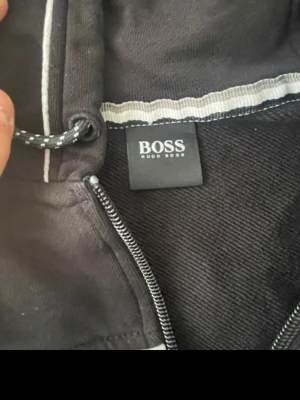 Svart hoodie från Hugo Boss - Snygg svart hoodie från Hugo Boss med dragkedja framtill och justerbar huva med snören. Klassisk design med diskret logga på insidan och detaljer i metall på dragkedjan. Perfekt för en stilren och avslappnad look.