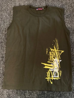 Grön ärmlös topp från Billabong - Cool grön ärmlös topp från Billabong med gul och vit grafisk text på framsidan. Perfekt för en avslappnad stil och passar bra till jeans eller shorts. Rund halsringning och mjukt material.