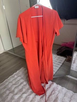 Orange långklänning med knytband - Snygg orange långklänning med korta ärmar och knytband i midjan. Klänningen har en enkel och stilren design, perfekt för sommardagar. Tillverkad i ett lätt och luftigt material som känns skönt mot huden.