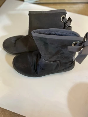 Grå boots från Xti med rosett - Säljer ett par grå boots från Xti med snygg rosettdetalj baktill. Skorna är i mockaimitation och har en platt sula med grovt mönster undertill. Perfekta för höst och vinter när du vill ha både stil och komfort.