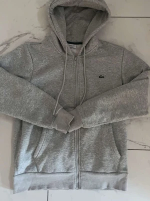 Grå zip hoodie från Lacoste - Snygg grå hoodie från Lacoste med dragkedja framtill och klassisk krokodil-logga på bröstet. Tröjan har huva med dragsko och två fickor framtill. Perfekt för en chill och sportig look. Tillverkad i mjukt sweatshirtmaterial.