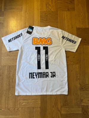 Vit Santos Neymar Jr matchtröja - Säljer en vit Santos matchtröja från Nike med Neymar Jr och nummer 11 på ryggen. Tröjan har tryckta sponsorer som BMG, Netshoes och Seara samt klubbmärke på bröstet. Tillverkad i lätt och ventilerande material, perfekt för fotboll eller träning.