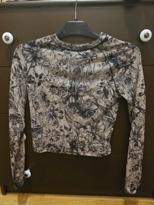 Blommig mesh topp från Bershka - Säljer en tunn och stretchig mesh topp från Bershka i beige med svart blommigt mönster. Toppen är långärmad och har en tight passform, perfekt att styla med jeans eller kjol.