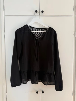 Svart blus med spetsdetalj - Snygg svart blus med långa ärmar och volang nedtill. Blusen har en unik spetsdetalj i ryggen och knytband vid halsen. Tillverkad i ett tunt, lätt material som ger ett luftigt intryck. Perfekt för dig som gillar detaljer och stilren design. Köpt på vila 