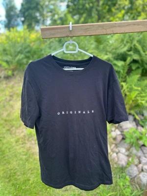 Svart t-shirt från Jack & Jones - Snygg svart t-shirt från Jack & Jones med texten 'ORIGINALS' tryckt i vitt på bröstet. Klassisk passform med rund hals och korta ärmar. Perfekt basplagg till garderoben och enkel att matcha med allt.
