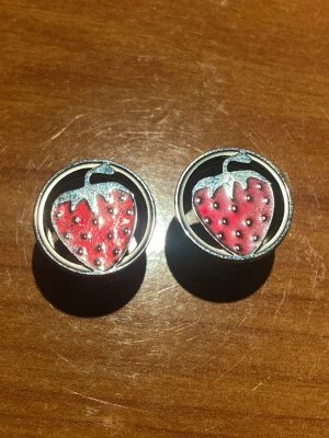 Runda örhängen med jordgubbsmotiv - Tunnlar med super söta jordgubbar. Diameter är 14mm och det finns gummiband till om man vill ha. Köpta men aldrig använda. Säljer båda för 50kr eller en för 30kr.