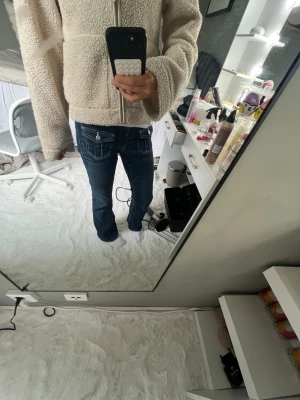 Mörkblå jeans med fickdetaljer från Gina Tricot  - Säljer ett par mörkblå bootcut jeans med snygga kontrastsömmar och dekorativa fickor med lock och knapp bak. Jeansen har låg midja och klassisk femficksdesign. Perfekta för dig som gillar en retro och avslappnad stil.